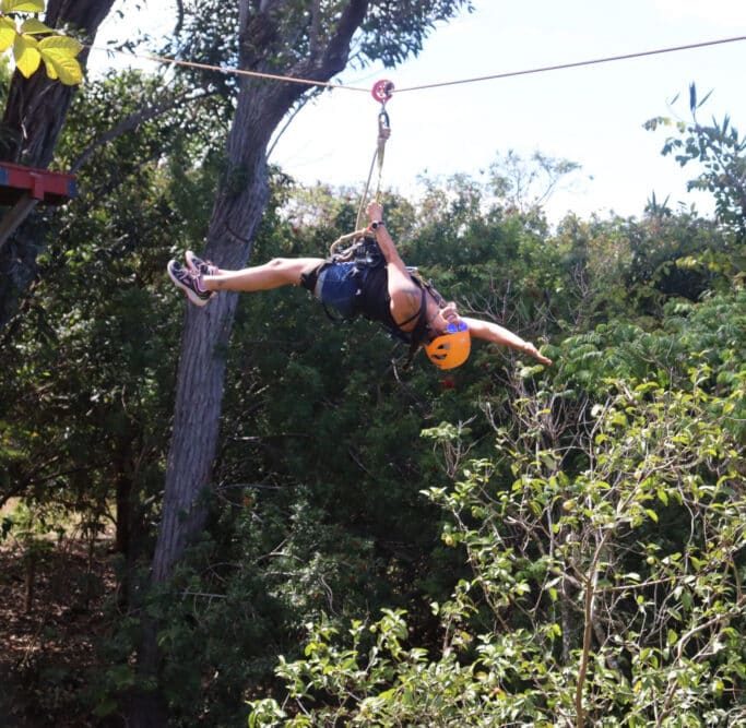 A person wearing a helmet and harness hangs sideways from a zipline above green trees, reaching one arm out while suspended in mid-air on a sunny day.