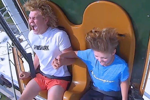 Two young people hold hands and scream with excitement while riding a thrill ride, their hair blown back by the wind. They are wearing casual shorts and T-shirts and are secured in their seats by safety harnesses.