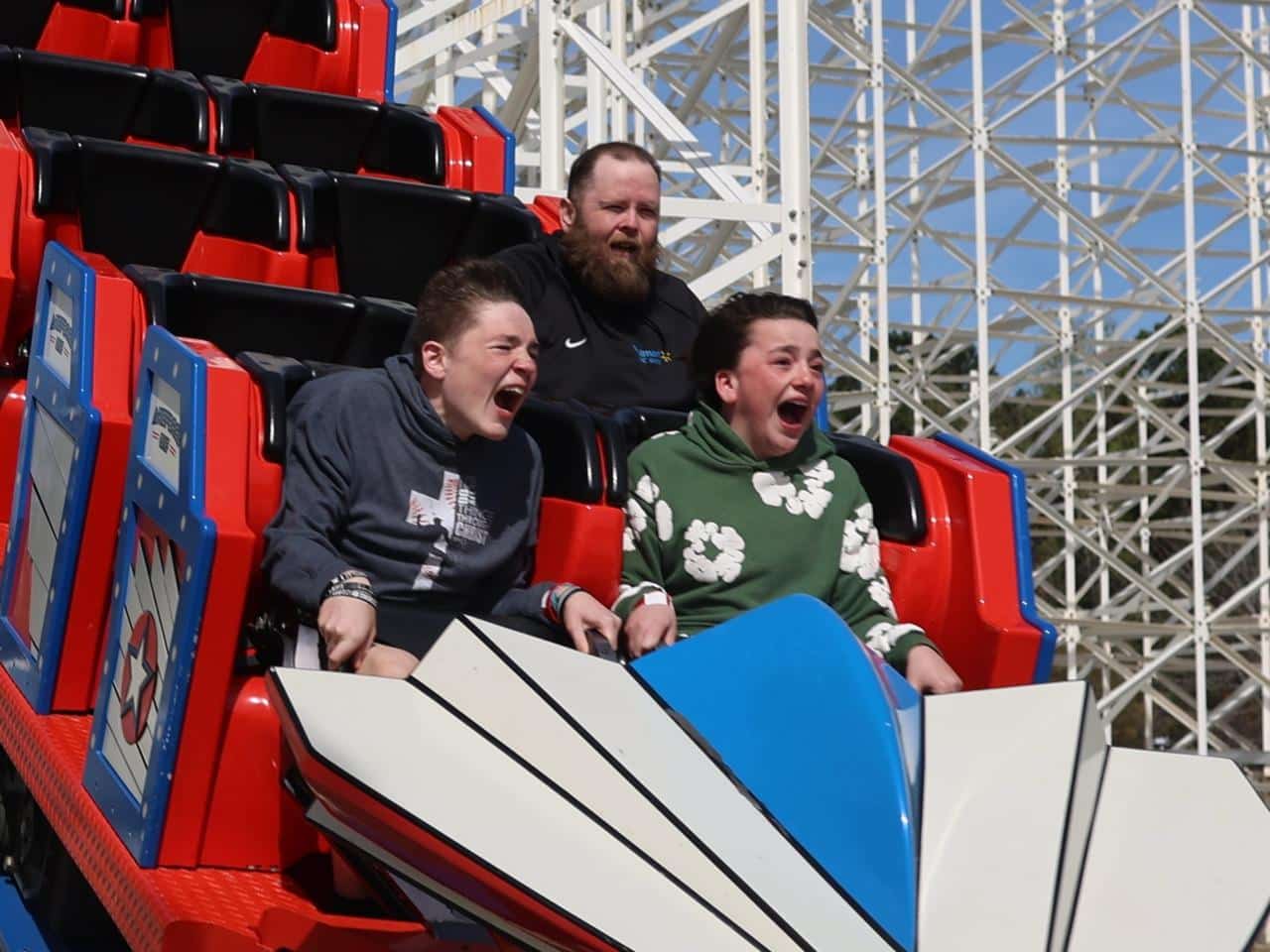 Three people ride a red roller coaster, two in front are screaming with excitement, and one behind looks calm. The coaster’s metal structure is visible in the background.