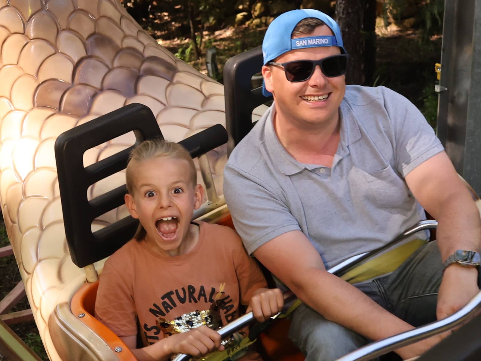 A smiling man and an excited child ride together on a roller coaster. The child has a surprised expression, and both are holding onto the safety bar. Sunlight shines on them, and trees are visible in the background.