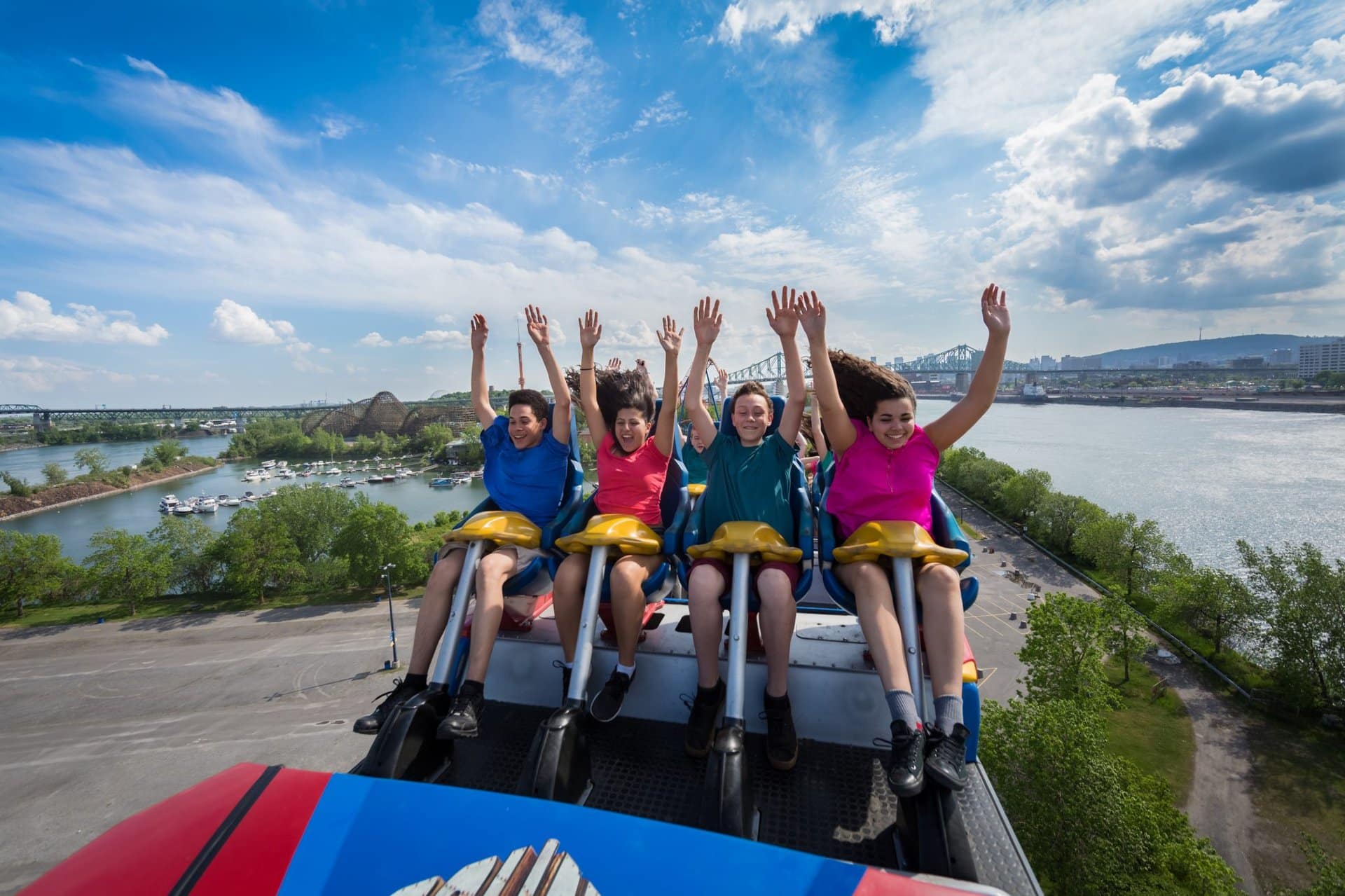 Four people ride a roller coaster with their hands up, smiling and excited. The coaster is at a peak, with a scenic view of water, trees, and a partly cloudy sky in the background.