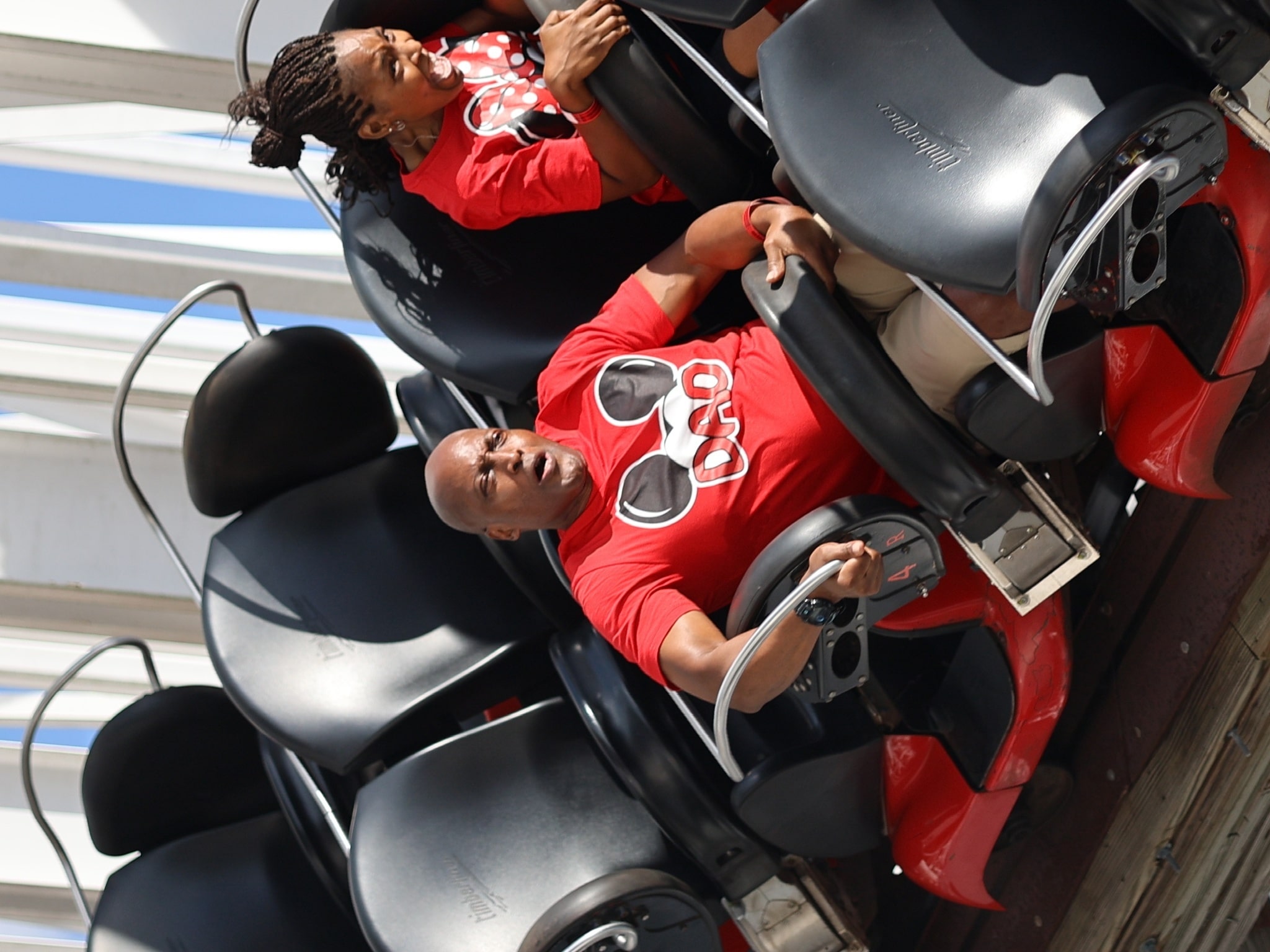 Two people in matching red shirts ride a roller coaster at the amusement park, gripping the safety bars and smiling as the car tilts, with empty seats visible beside them.