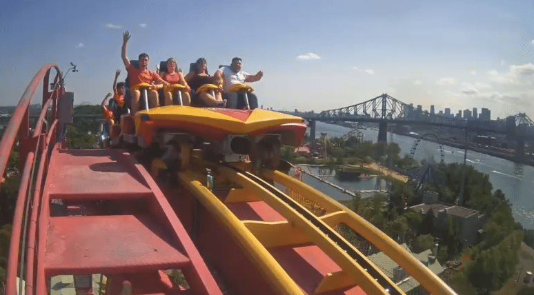 People ride a roller coaster at the top of a hill, with hands raised, overlooking a city skyline, river, and bridge under a clear blue sky.