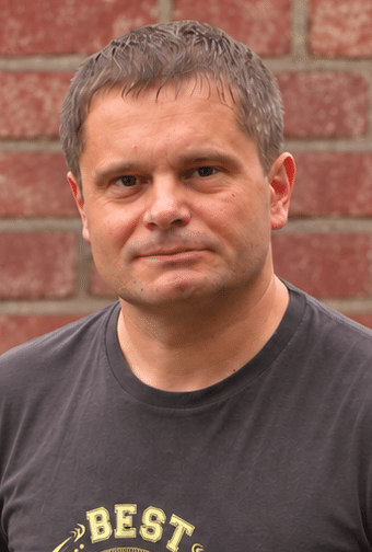 A man with short brown hair stands in front of a brick wall, wearing a dark T-shirt with yellow writing. He looks directly at the camera with a neutral expression.