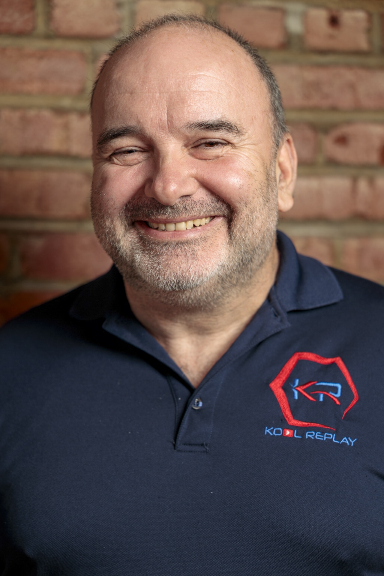 A smiling man with a beard wearing a navy blue polo shirt with a red and blue “Kool Replay” logo stands in front of a brick wall.