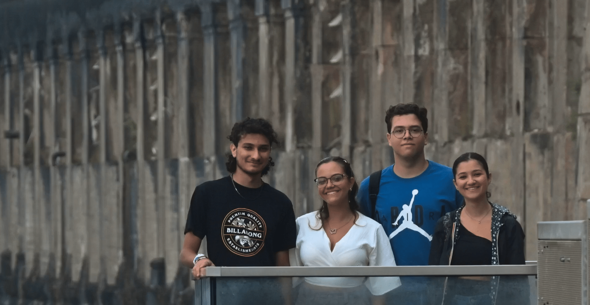 Four young adults stand and smile at the camera in front of a tall stone wall. Two men are on the left, and two women are on the right. One man wears glasses and a blue shirt with a backpack.