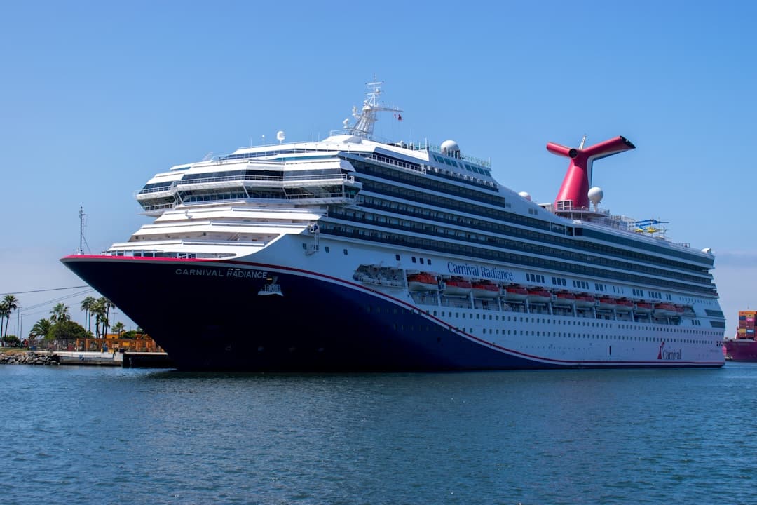 A large white and blue cruise ship named Carnival Radiance, part of one of the world’s leading cruise lines, is docked at a port on a sunny day, with clear skies and calm water.