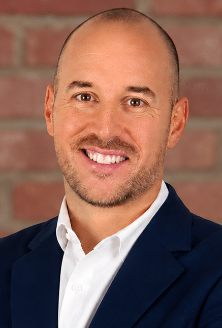 A smiling man with a shaved head and short beard, wearing a white shirt and dark blue blazer, stands in front of a brick wall background.