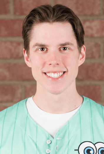 A young man with short brown hair smiles at the camera. He is wearing a light green shirt with a white undershirt. The background is a brick wall.