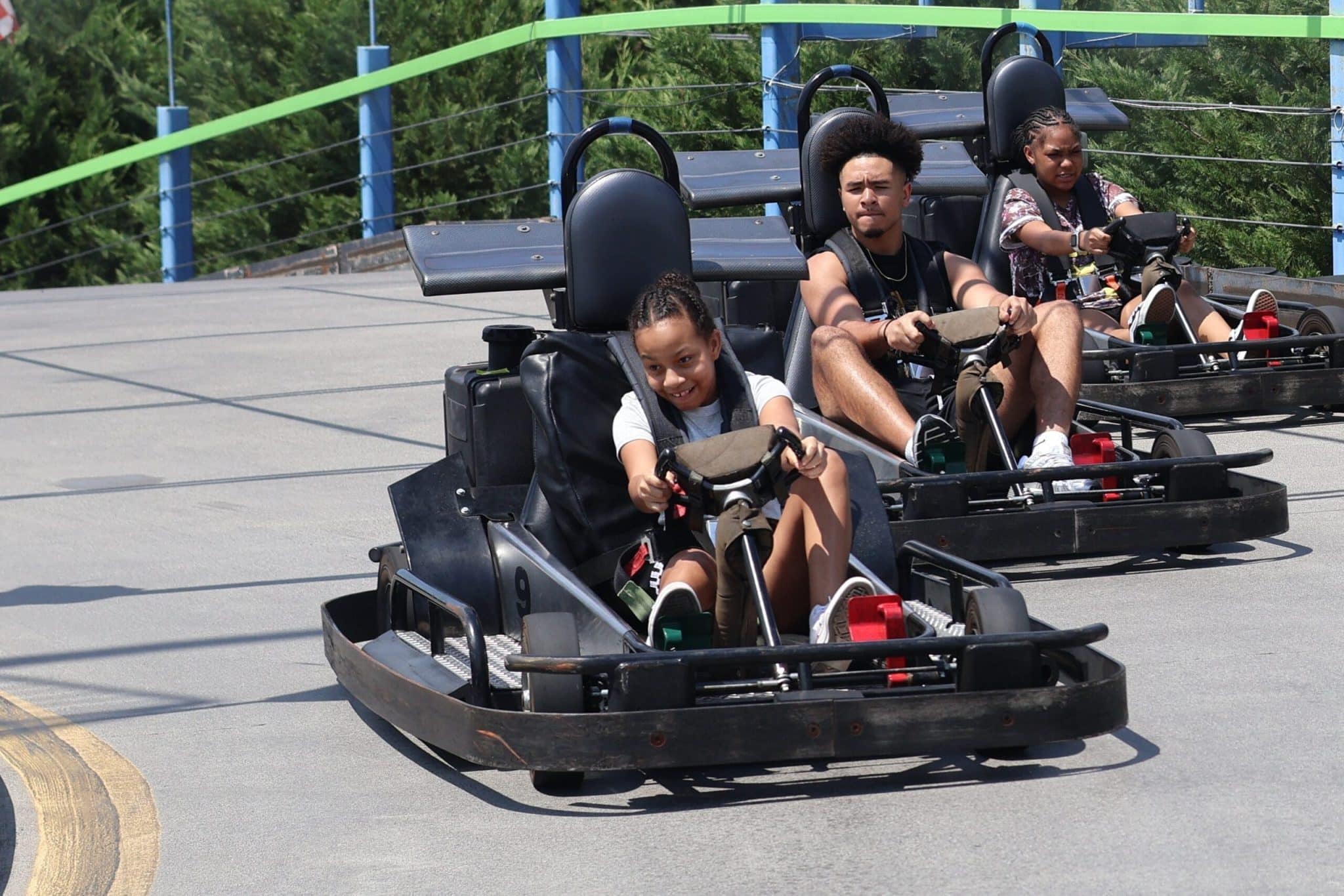 Three people ride go-karts on an outdoor track. A young girl in the front smiles while steering, with two other racers, a man and a child, following behind her. Green trees and safety rails are in the background.