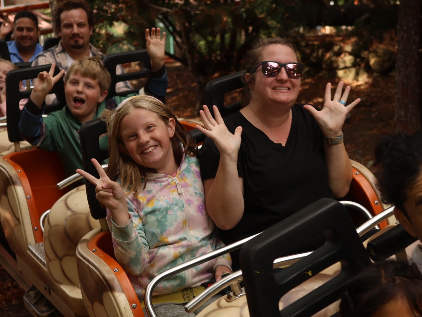 A group of people on a roller coaster, smiling and raising their hands. A woman wearing sunglasses and a girl in a pastel hoodie are in front, with trees and sunlight in the background.