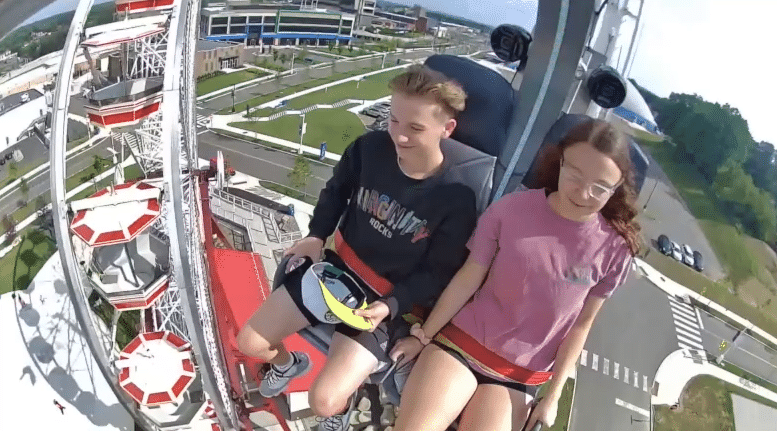 Two young people sit side by side on an amusement park ride, high above the ground, secured with red harnesses. With one tower rising in the background and hinting at even more thrills among the park’s four rides, the scene also features a view of roads, buildings, and greenery below.