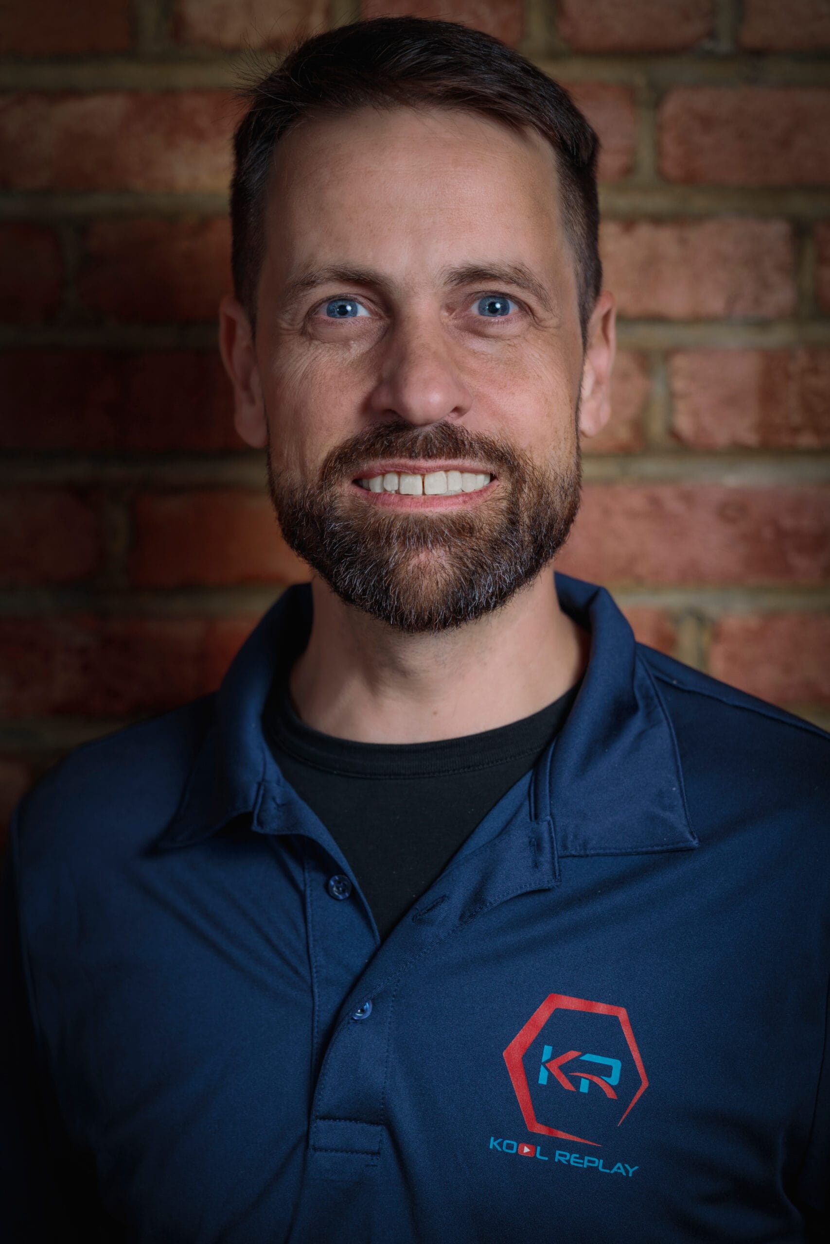 A smiling man with a beard wearing a dark blue polo shirt with a “KOOL REPLAY” logo stands in front of a brick wall.