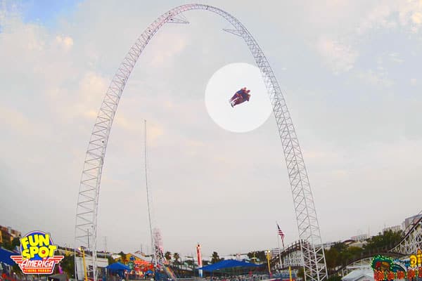 Two people are riding a large swing ride, suspended high in the air between two tall metal towers at an amusement park, with the Fun Spot America logo visible in the corner and excitedly anticipating their next thrill as they check ticket availability for more ride tickets.