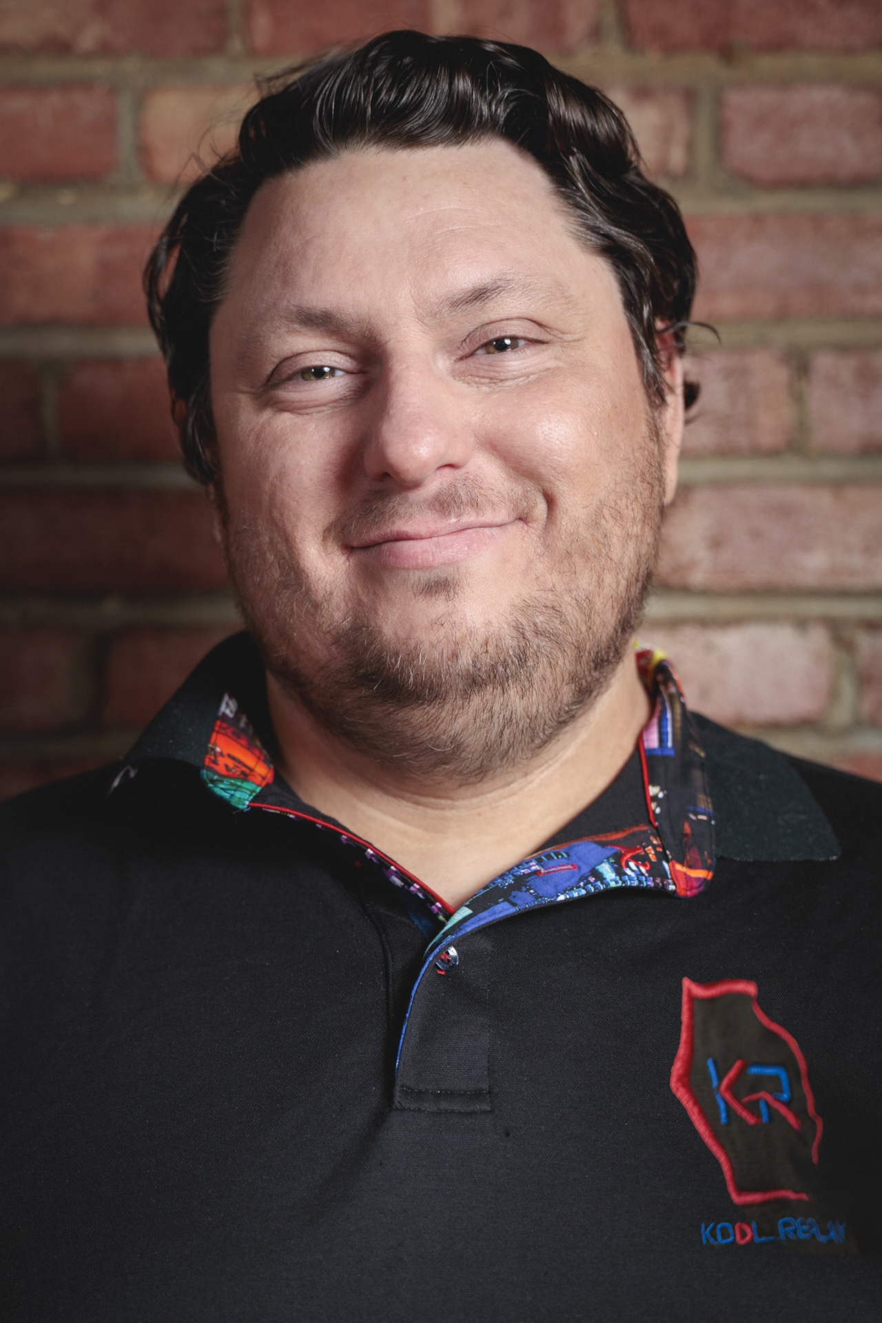 A man with short dark hair and a beard smiles at the camera, standing in front of a brick wall. He is wearing a black shirt with colorful trim and a red and blue embroidered logo on the chest.