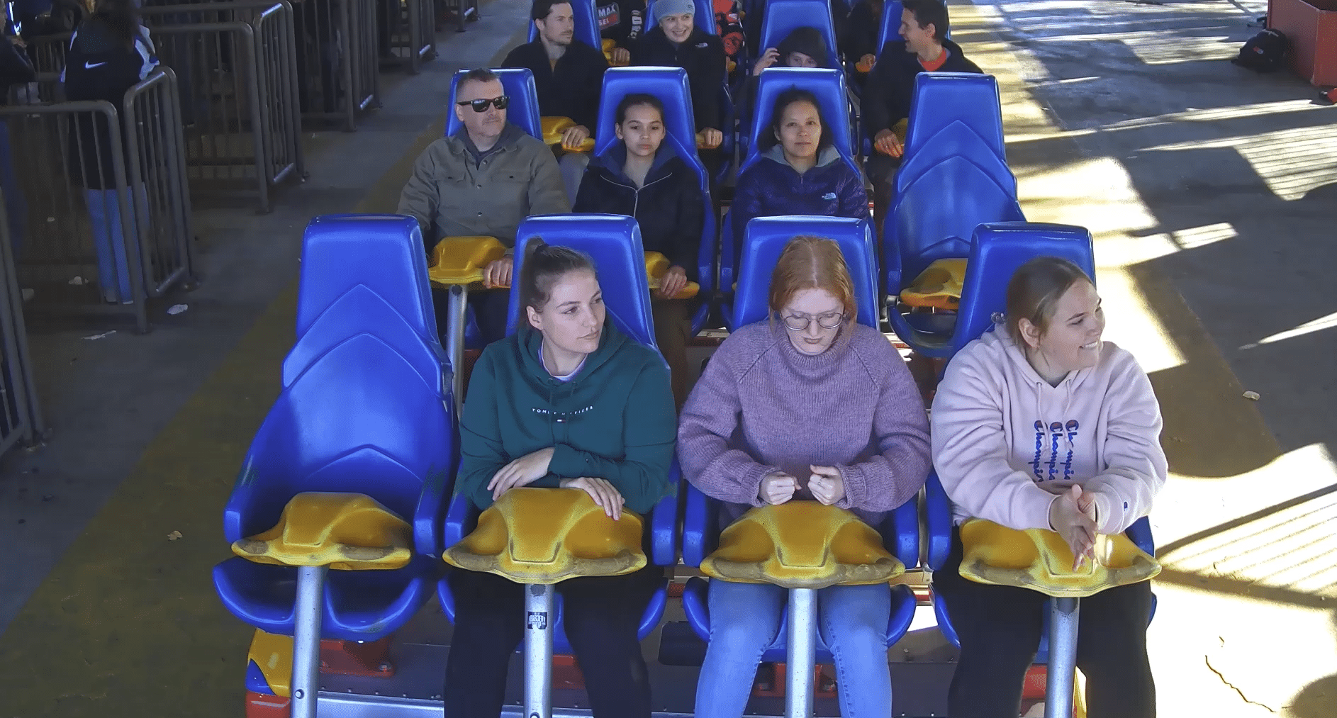Nine people sit in blue and yellow seats on a roller coaster, waiting for the ride to start. Four are in the front row, and five are behind them. Most are looking ahead, while some look to the side.
