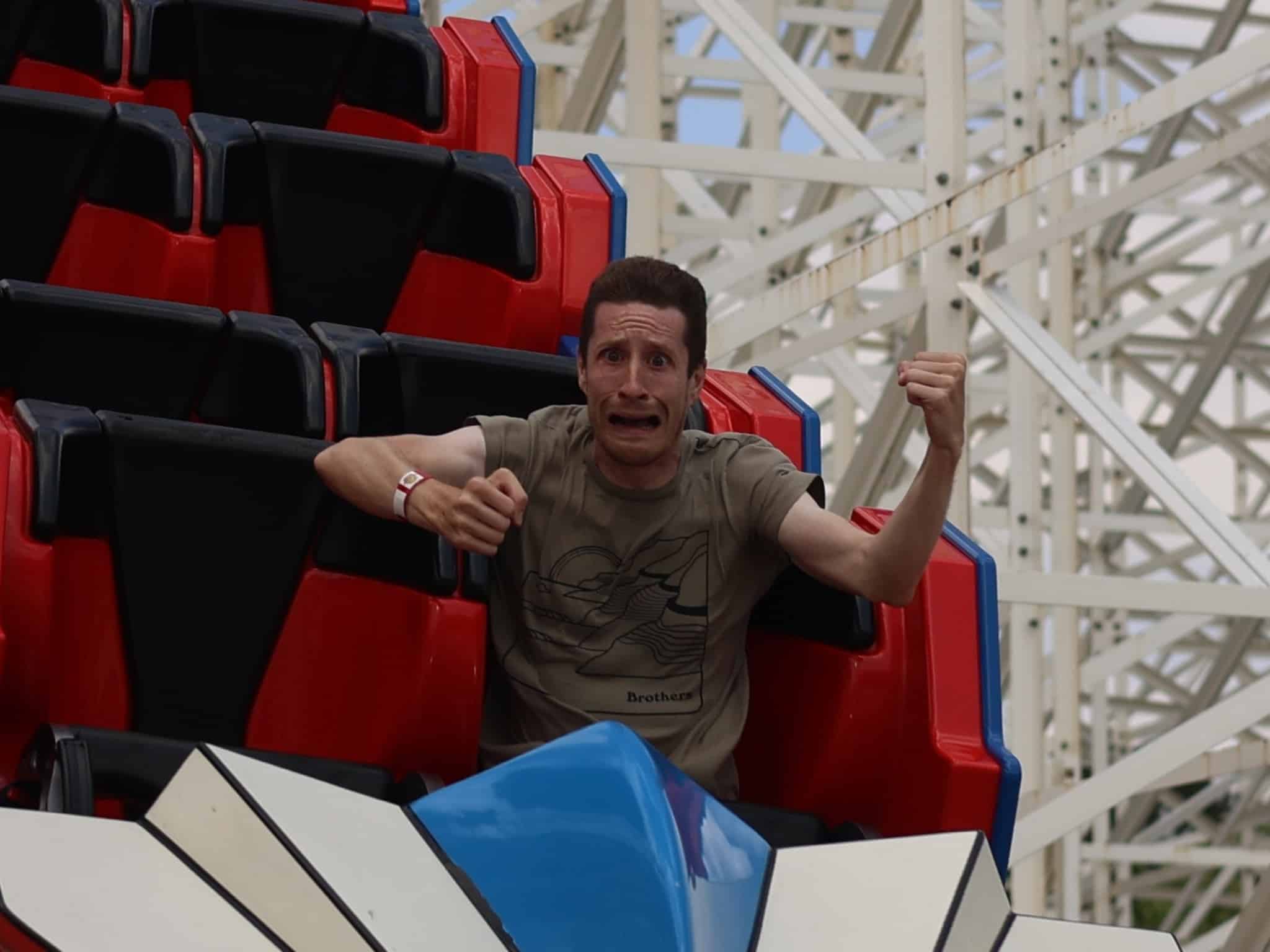 A person with a scared expression clutches the side of a roller coaster car while riding through an Amusement Park, surrounded by red and black seats and white support beams in the background.