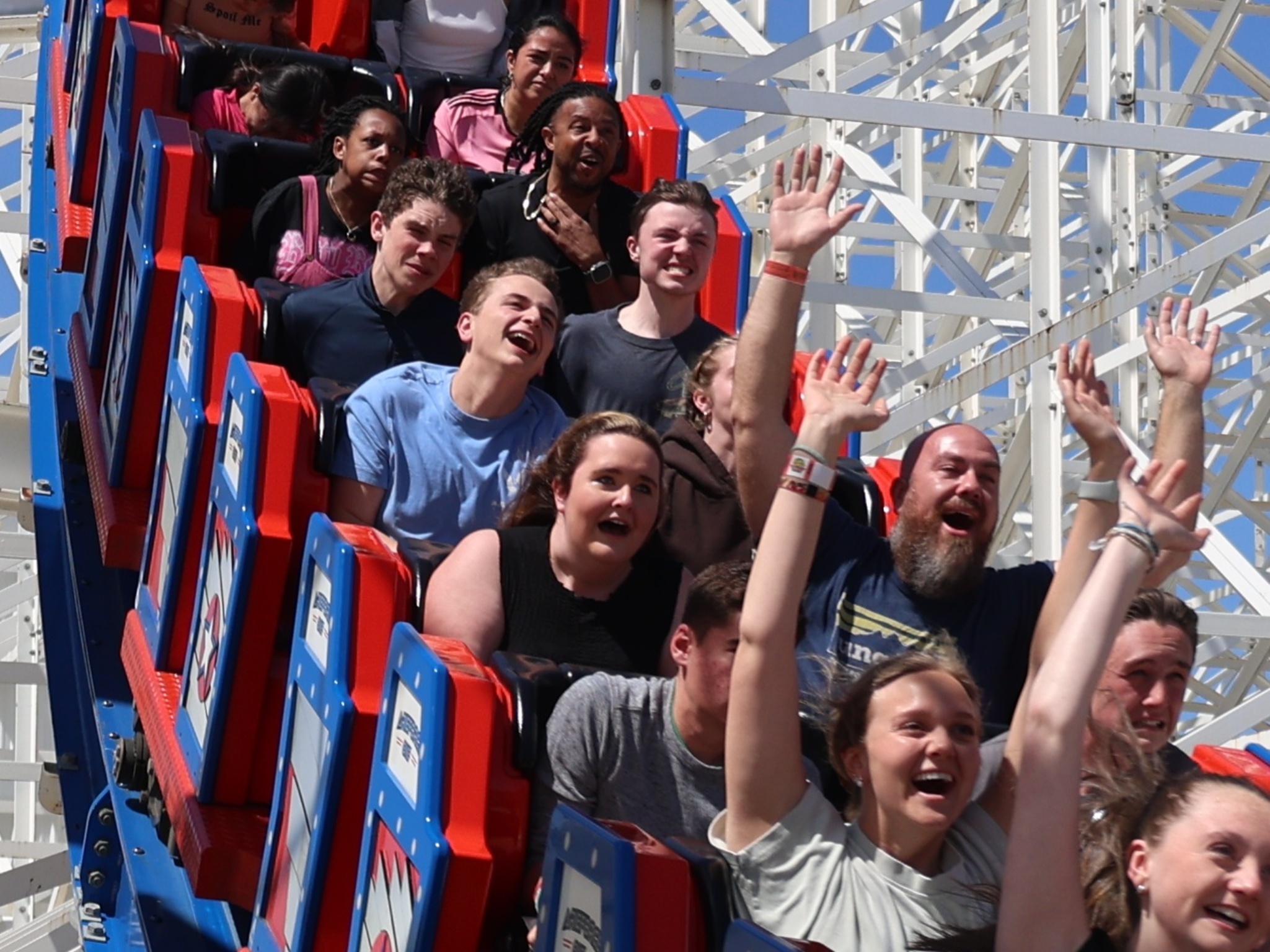 People of various ages ride a roller coaster at one of the most thrilling amusement parks, some with hands up and excited expressions, others looking nervous. The coaster features red and blue seats, and white support beams are visible in the background.