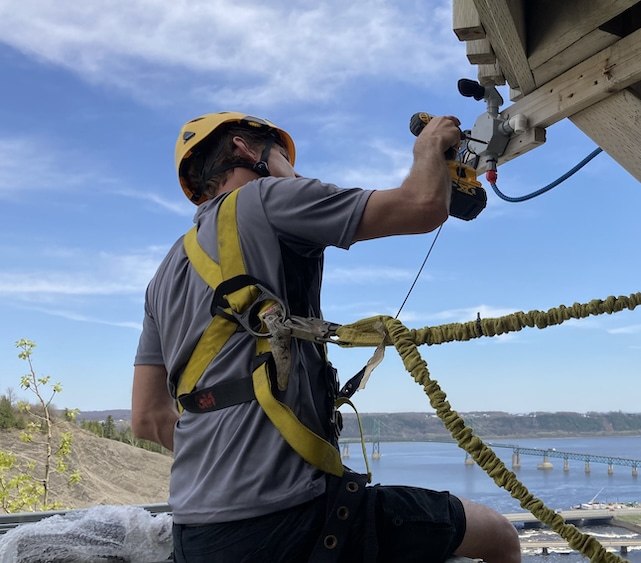 A worker wearing a safety harness and helmet uses a power drill to install equipment on a wooden structure, with a river and bridge visible in the background.