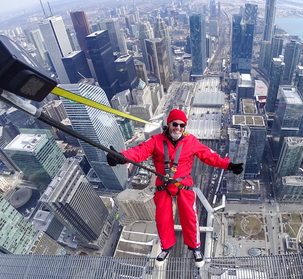 A person in red safety gear, harnessed to a cable, stands on a ledge high above a city skyline, smiling with arms outstretched. Skyscrapers and streets are visible far below.