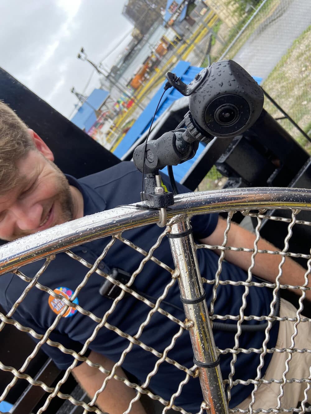 A man in a navy shirt stands outside near a metal net, with a small camera attached to the net. The scene is wet with raindrops visible on the equipment.