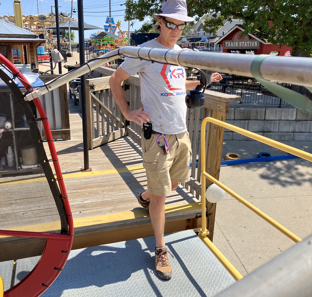 A man in a hat and sunglasses stands on a wooden platform near a train ride at an amusement park, with trees and buildings visible in the background on a sunny day.
