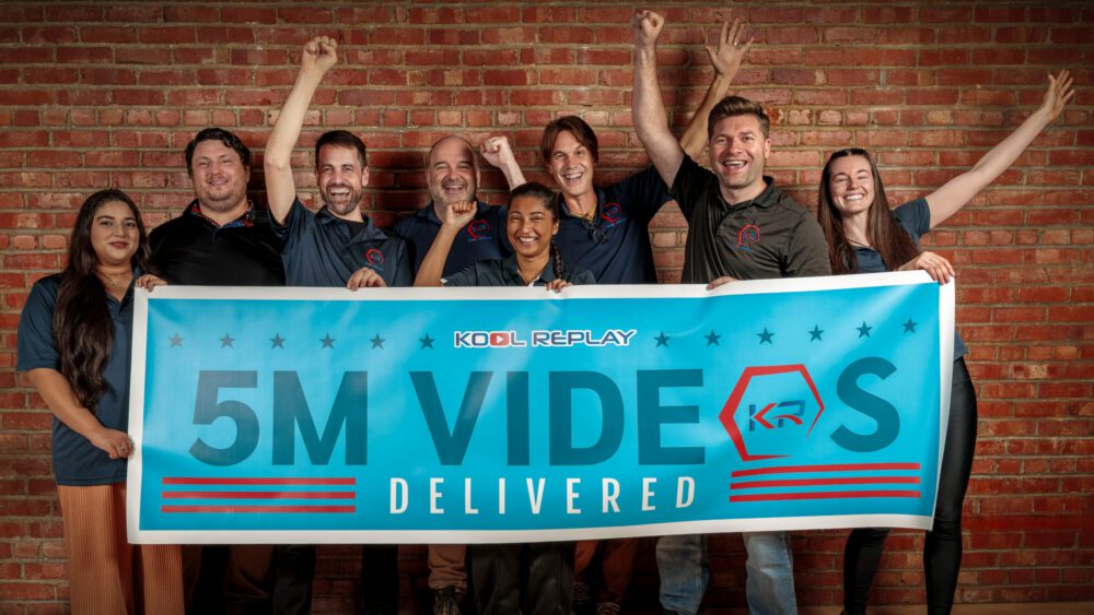 Eight people stand smiling in front of a brick wall, holding a banner that says “KOOL REPLAY 5M VIDEOS DELIVERED.” Some are raising their arms in celebration.