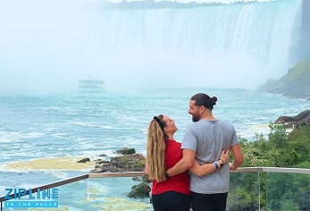 A couple stands on a viewing platform, smiling at each other with arms around one another, overlooking Niagara Falls. A tour boat is visible in the mist below.