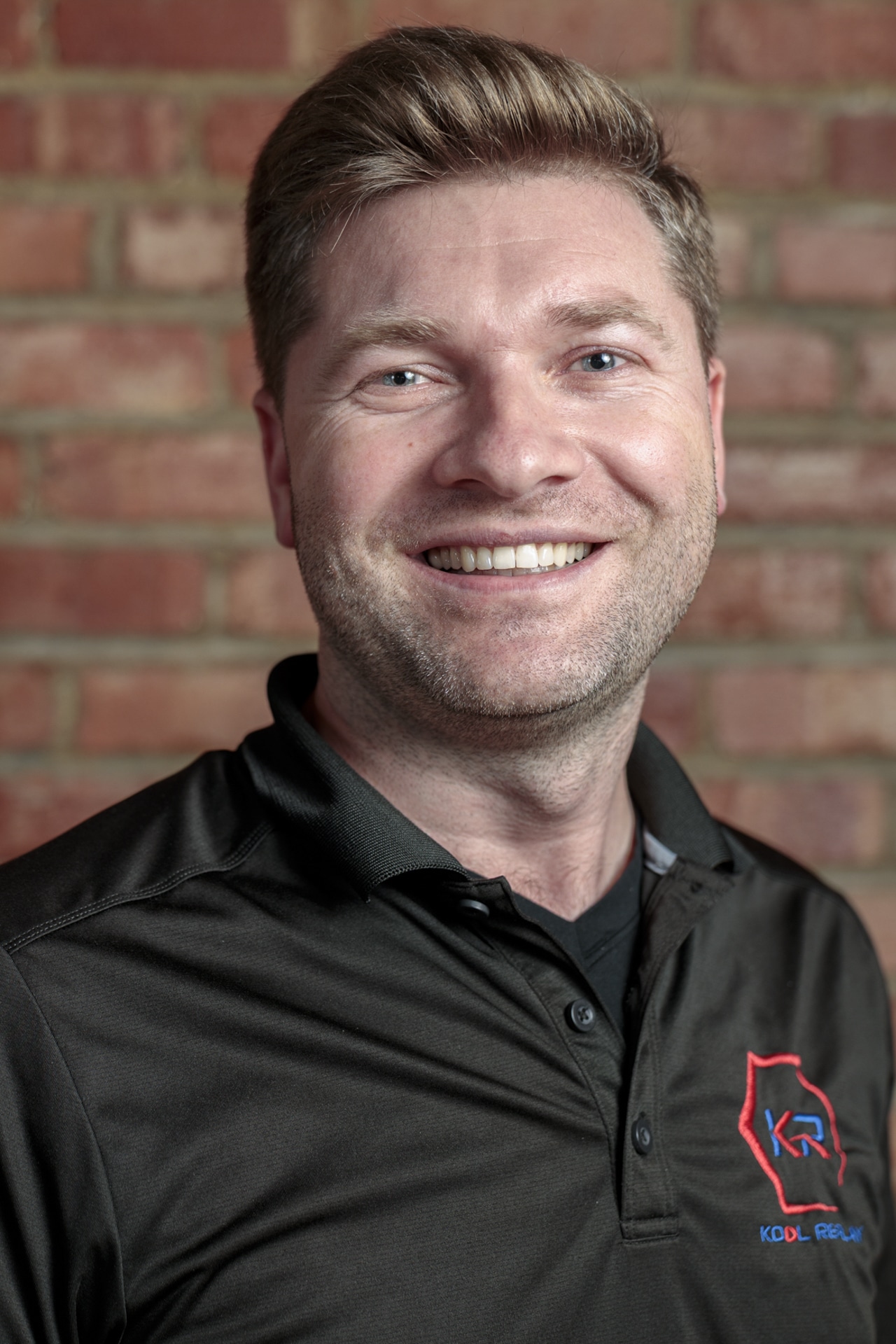 Smiling man with short light brown hair wearing a black polo shirt with a red and blue logo, standing in front of a brick wall.