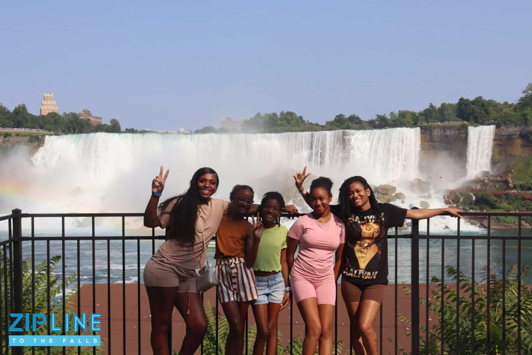 Five smiling girls pose in front of a black fence with a large waterfall and a rainbow in the background. The sky is blue, and greenery surrounds the scene. The “ZIPLINE TO THE FALLS” logo appears in the bottom left corner, highlighting an unforgettable Zipline Ride experience against this stunning natural backdrop.