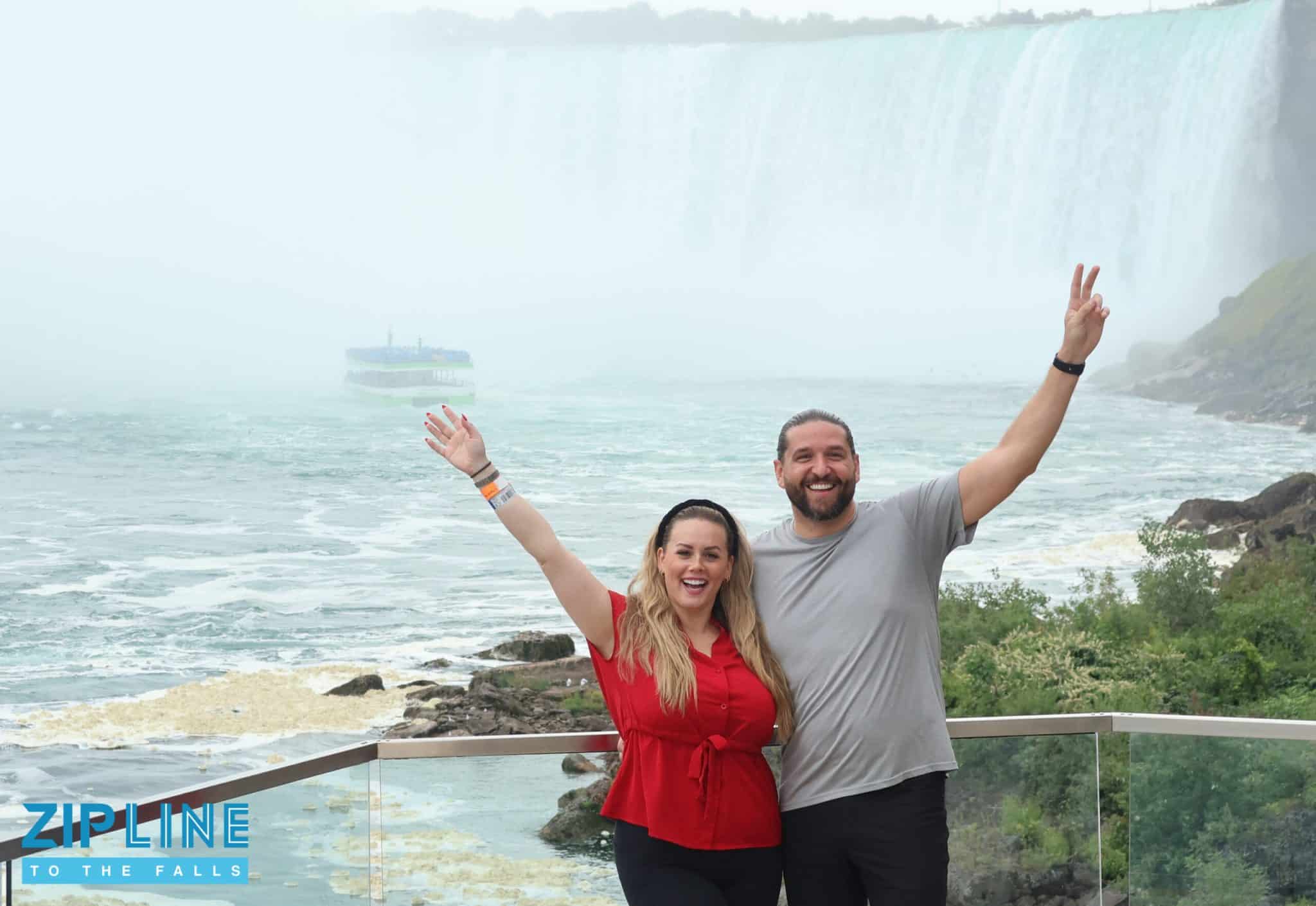 A smiling couple poses with arms raised in front of Niagara Falls, capturing the thrill of adventure tourism. Mist from the waterfall and a tour boat are visible in the background. “Zipline to the Falls” logo appears in the bottom left corner.