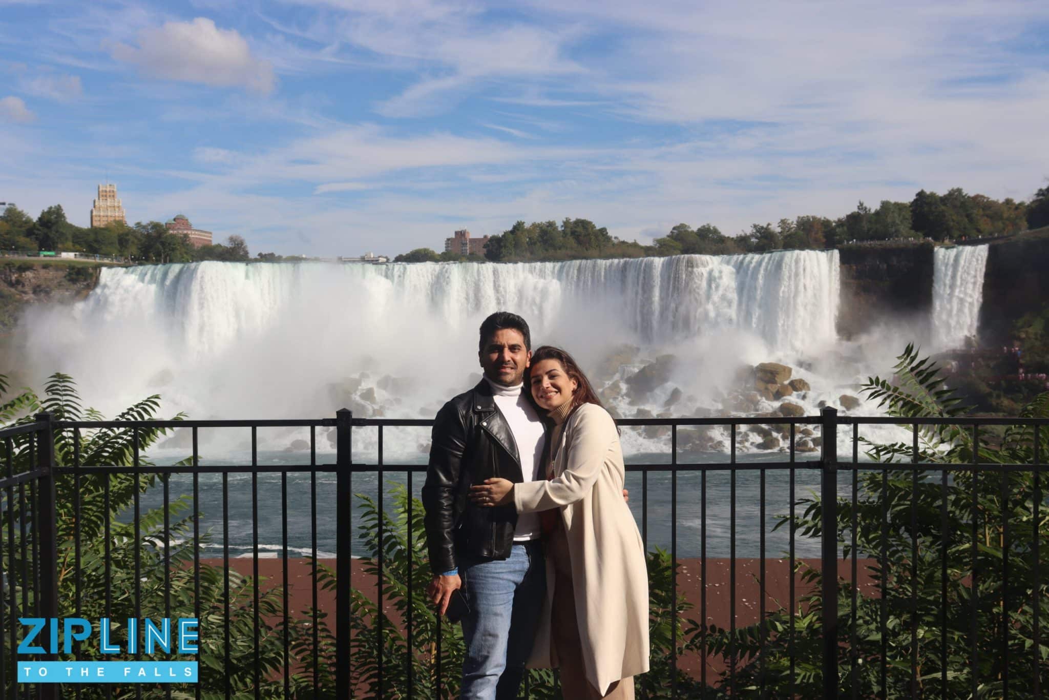 A couple poses together in front of Niagara Falls on a sunny day, standing by a black fence with greenery around. A “Zipline to the Falls” sign is visible in the lower left corner, inviting adventurous visitors to enjoy an exhilarating zipline ride with stunning views of the falls.