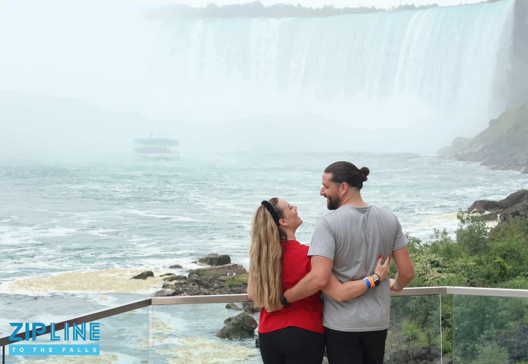 A couple stands on a viewing platform hugging, looking at each other, with Niagara Falls in the background. The excitement of the Zipline Ride brings them even closer together. “ZIPLINE TO THE FALLS” text is in the bottom left corner.
