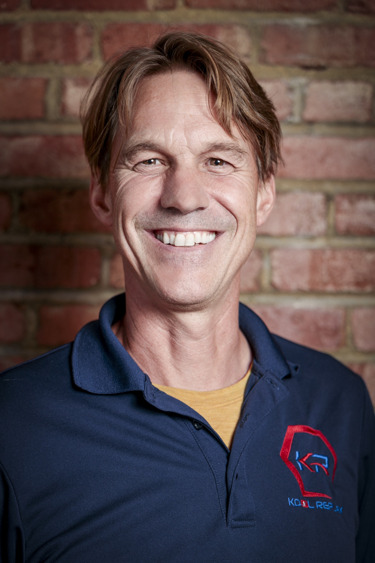 A smiling man with light brown hair wearing a blue polo shirt with a red and blue logo stands in front of a brick wall.