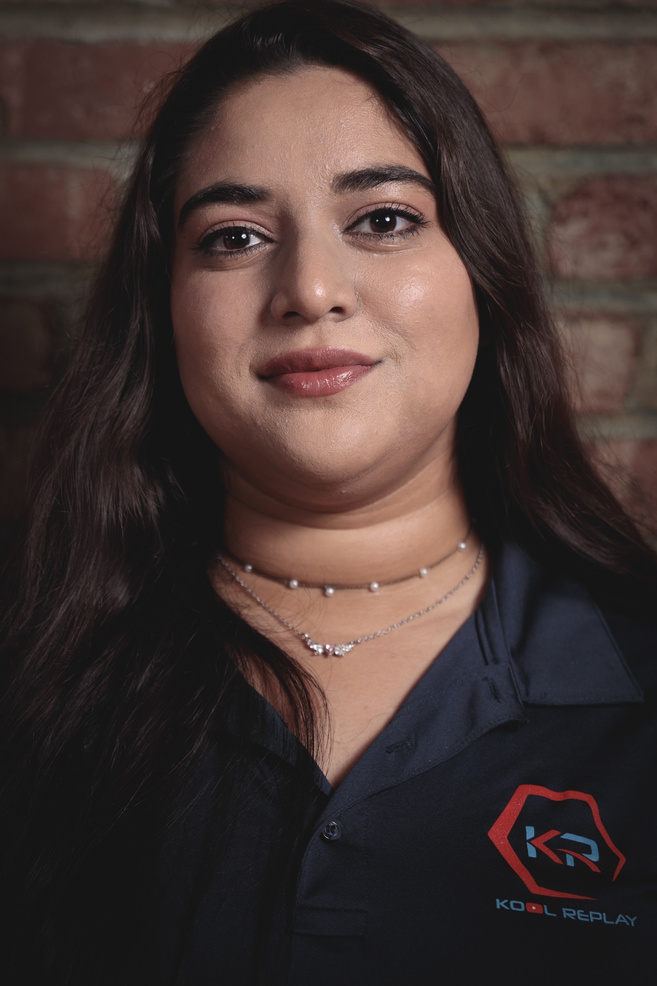 A woman with long dark hair, wearing a navy shirt with a “Kool Replay” logo and layered necklaces, stands in front of a brick wall, looking at the camera with a slight smile.