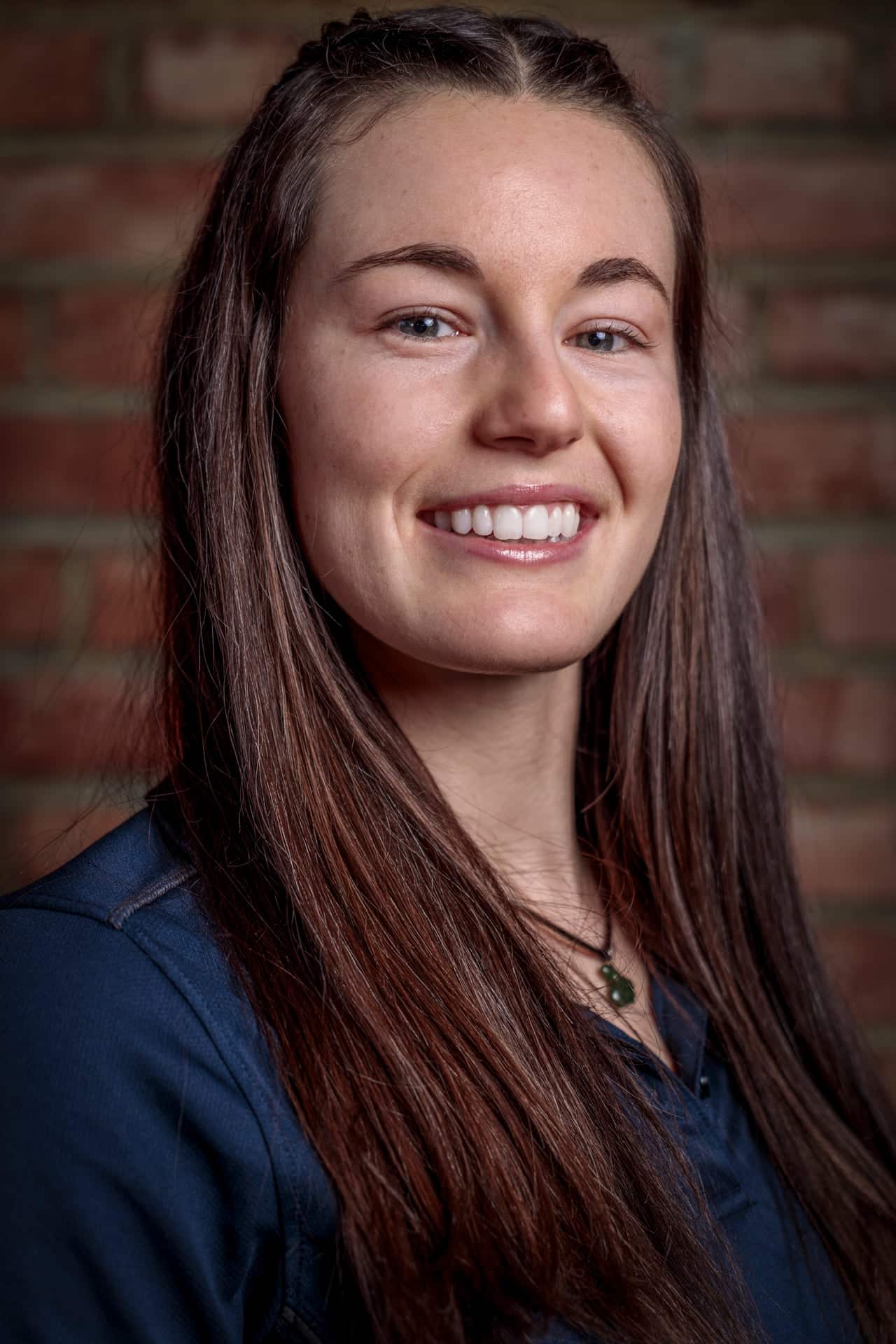 A woman with long brown hair and a blue shirt smiles in front of a brick wall background.