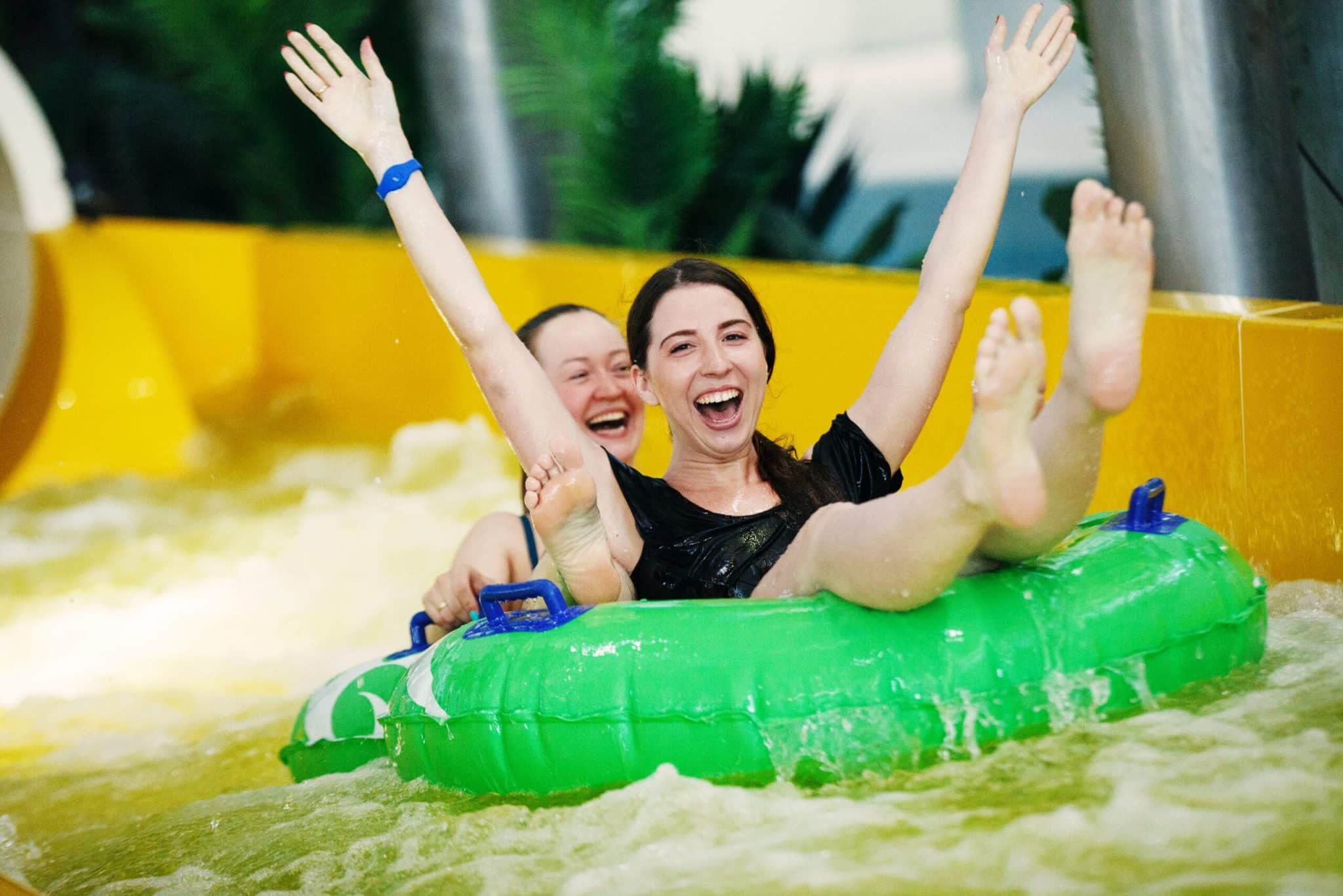 Two women laugh and raise their arms while riding together on a green inflatable tube down a water slide with yellow walls.