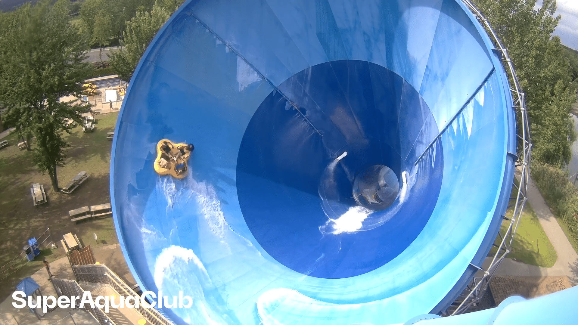 Two people ride a yellow inflatable tube inside a large blue funnel water slide at a water park. The SuperAquaClub logo appears in the bottom left corner. Trees and walkways are visible outside the slide.