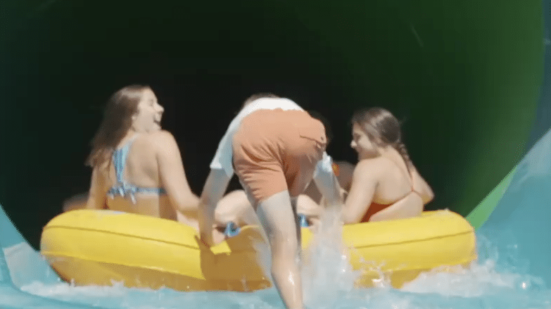 Three people on a yellow raft prepare to enter a dark water slide tunnel, with two laughing and facing forward as water splashes around them.