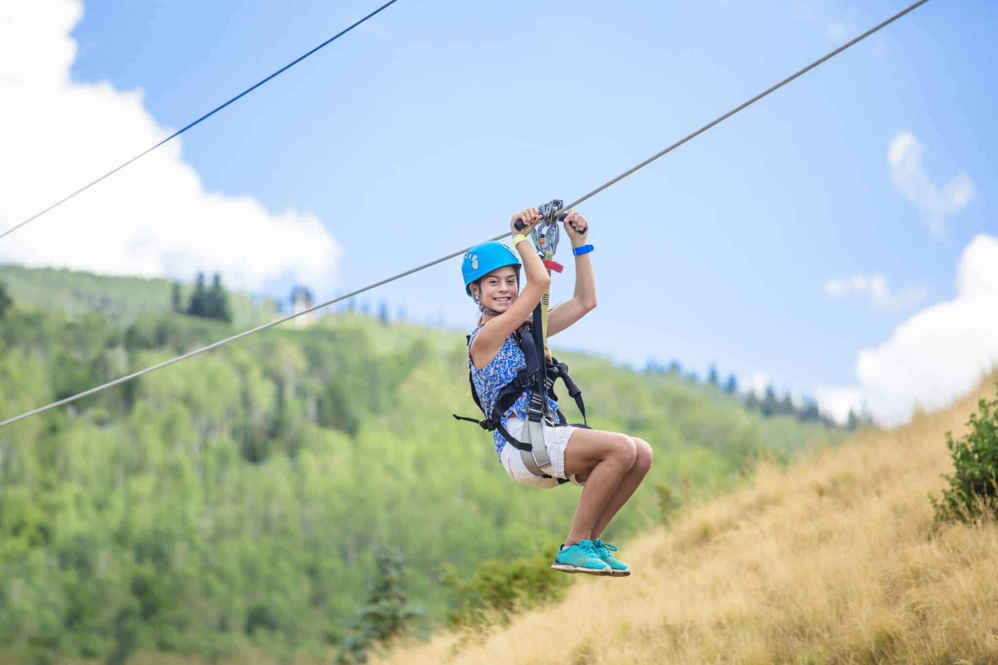 A smiling child in a blue helmet enjoys an exhilarating zipline ride over grassy hills and green trees on a sunny day, with a blue sky and clouds in the background.