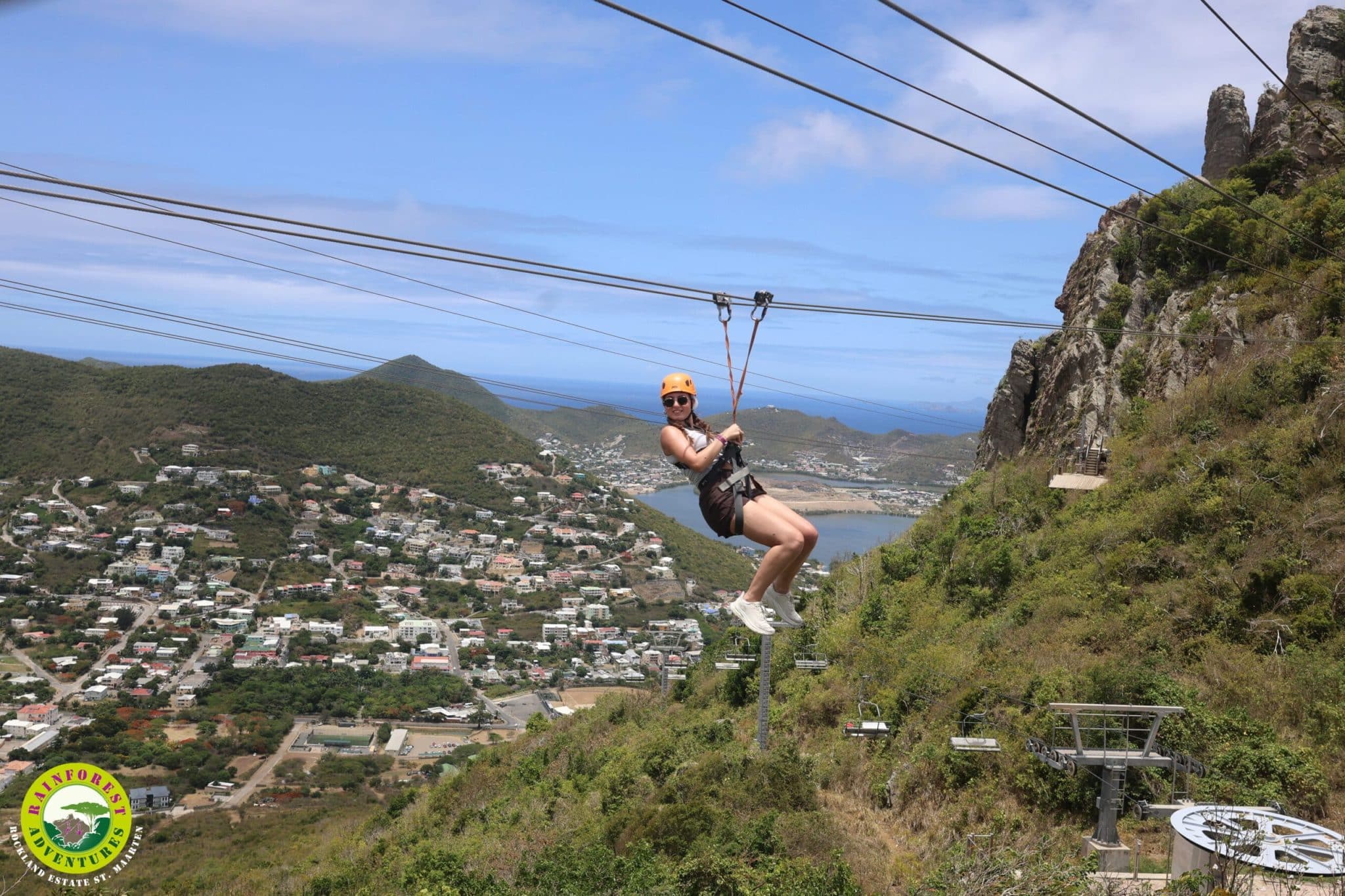 A person wearing a helmet and harness is enjoying an exhilarating Zipline Ride over a lush, hilly landscape with a town and water in the background under a partly cloudy sky.
