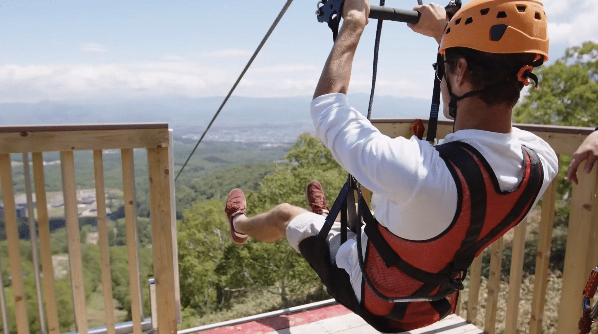 A person wearing a helmet and harness is about to enjoy a thrilling zipline ride from a wooden platform, overlooking a green landscape and distant mountains.