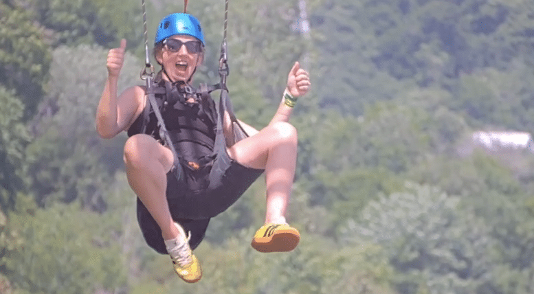 A person wearing a helmet and harness smiles and gives two thumbs up while enjoying a thrilling zipline ride outdoors above green trees.